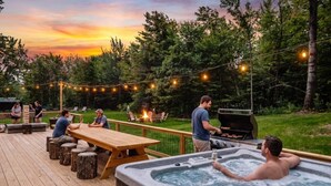 Una terraza con jacuzzi, mesas y sillas de madera, rodeada de árboles y luces colgantes.
