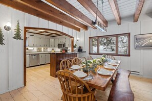A dining area with a wooden table set for six, a kitchen in the background, and a window with a snowy view.