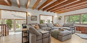 A spacious living room with a large sectional sofa, wooden beams, and large windows offering a view of the outdoors.