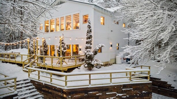Un edificio blanco con varias ventanas, una terraza de madera y árboles cubiertos de nieve.