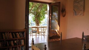 Ein Zimmer mit einem Holztisch, Stühlen und einem Bücherregal. Eine Tür führt in einen Außenbereich mit einem Baum und Blick zum Himmel.