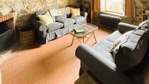 Ein rustikales Wohnzimmer mit freiliegenden Holzbalken, einer Steinwand, einem grauen Sofa, einem Korbtisch und einem Bücherregal.