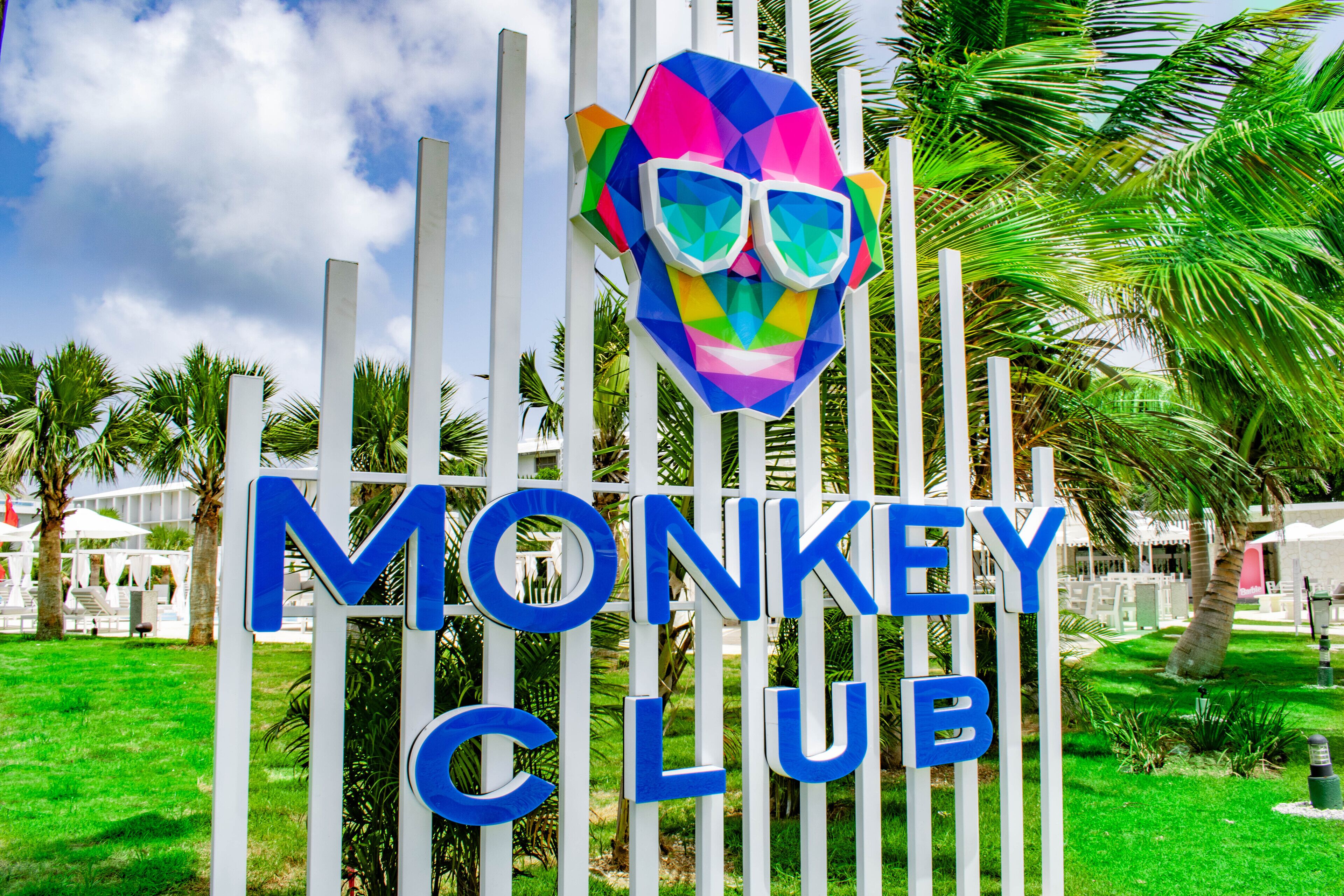 Sininen kyltti, jossa lukee suurilla kirjaimilla ”MONKEY CLUB”. Kyltin ympärillä on valkoisia, pystysuoria pylväitä ja taustalla näkyy palmuja.