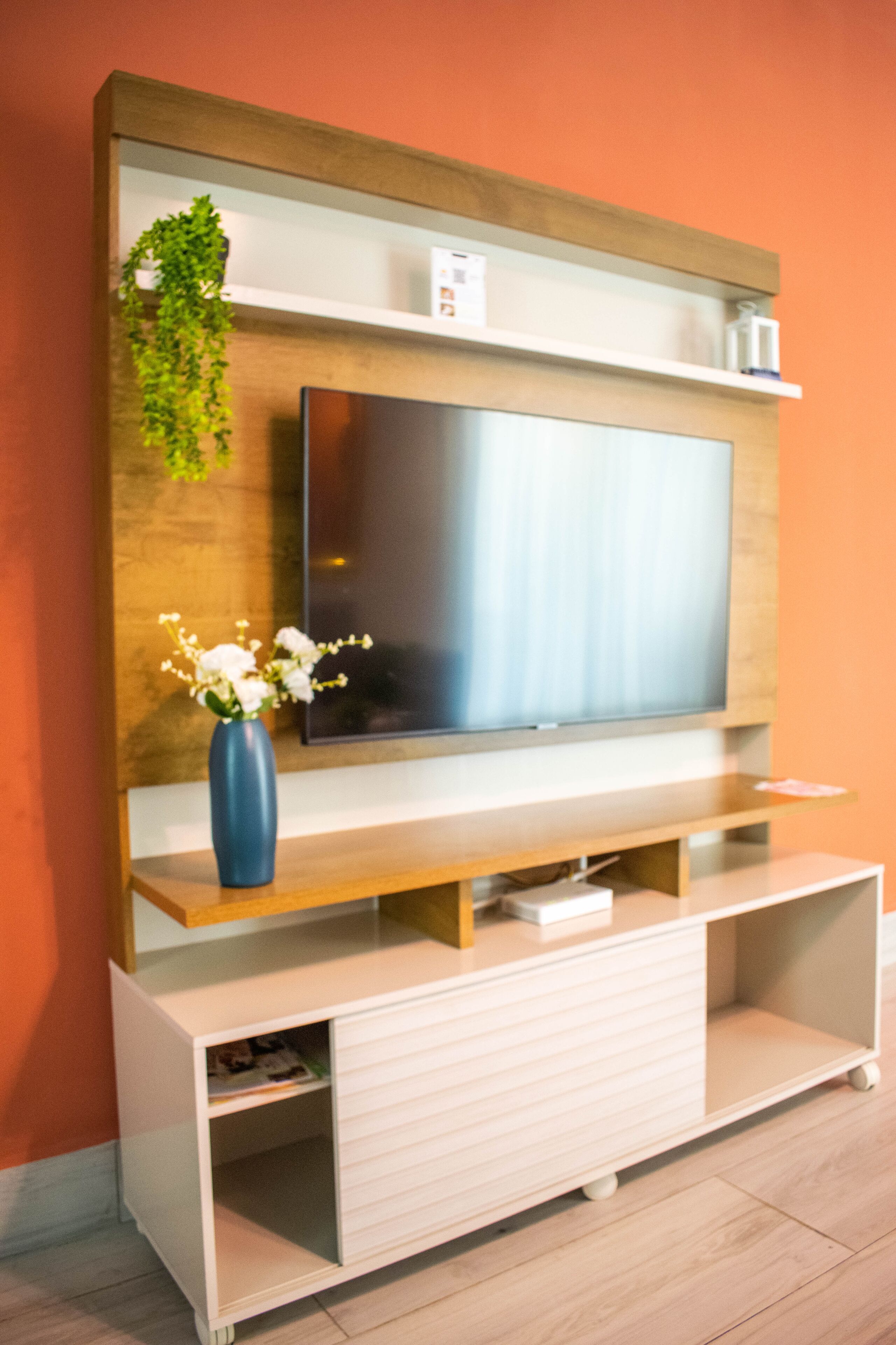 A modern living room with a flat-screen TV mounted on a wooden wall, a blue vase with white flowers on a wooden console, and a small shelf above the TV.