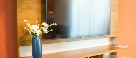 A modern living room with a flat-screen TV mounted on a wooden wall, a blue vase with white flowers on a wooden console, and a small shelf above the TV.