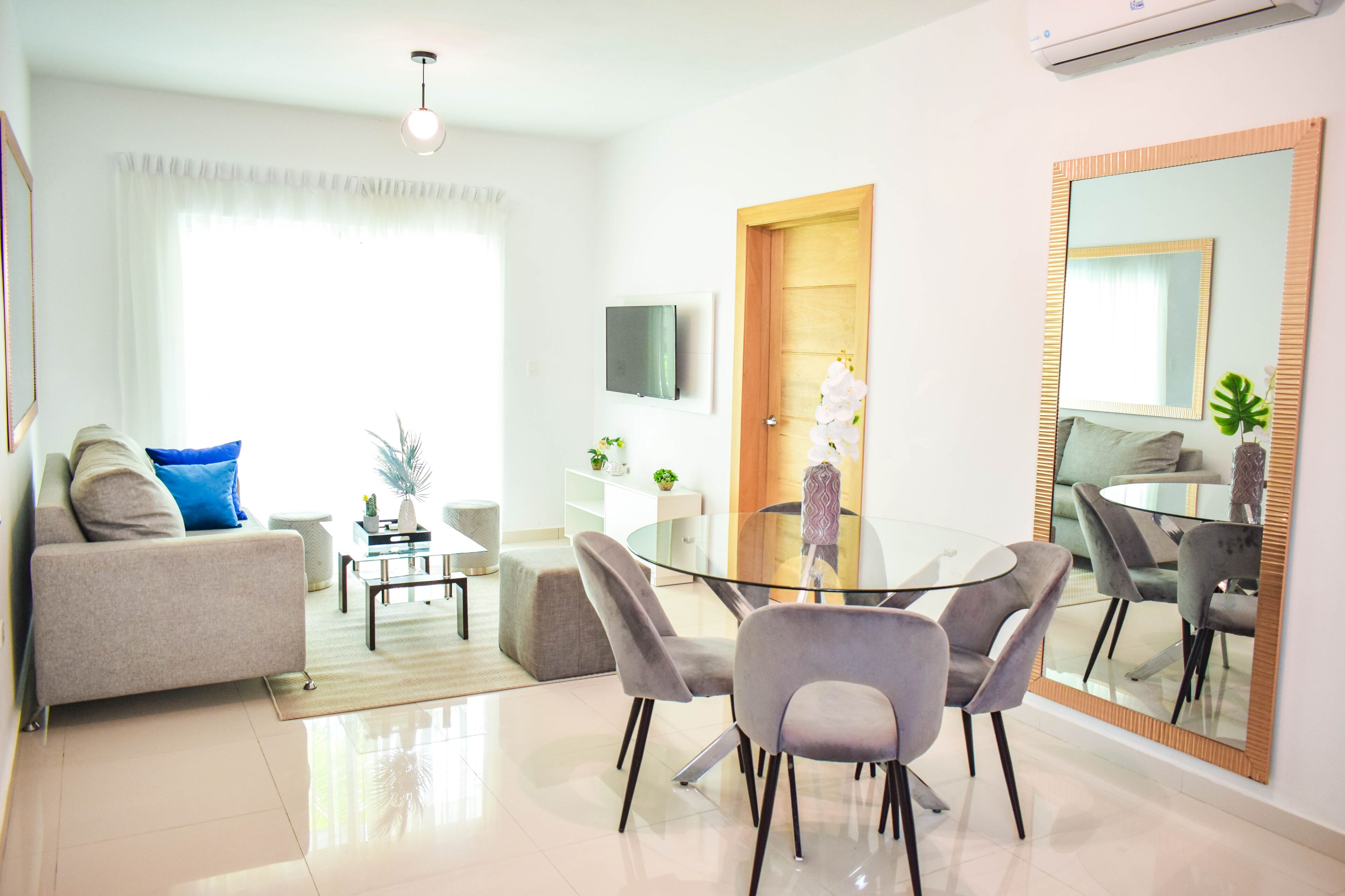 A modern living room with a glass dining table, grey sofas, and a large mirror.