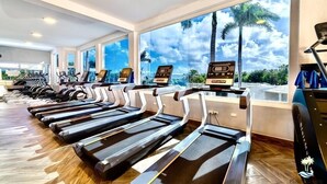 A gym with treadmills aligned along a wall with large windows overlooking palm trees and a cloudy sky. - Playa Palmera Beach Resort  (Punta Cana)