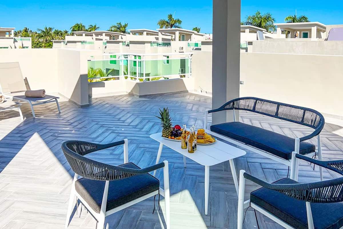 A rooftop terrace with a white table, two blue chairs, and a small sofa. There is a fruit bowl and bottles on the table.