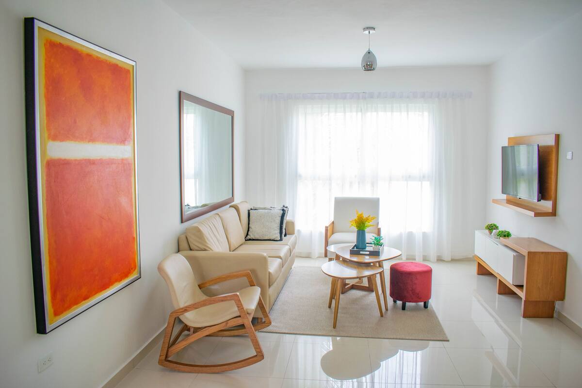 A modern living room with a beige sofa, a wooden rocking chair, a small round table with a vase, and a red ottoman.