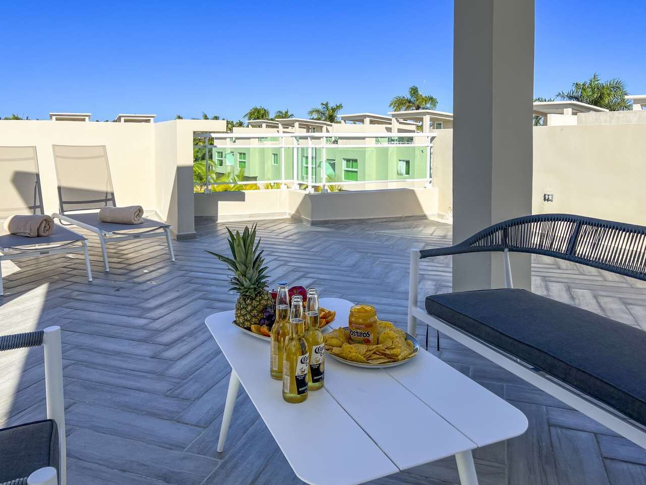 Een terras op het dak met een tafel gedekt voor drankjes en hapjes, zitplaatsen buiten en uitzicht op een gebouw met balkons.