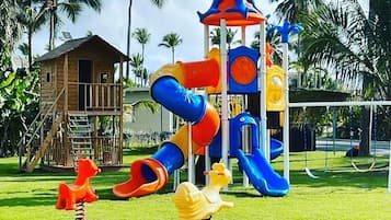 A playground with slides and swings, surrounded by palm trees and a wooden structure.
