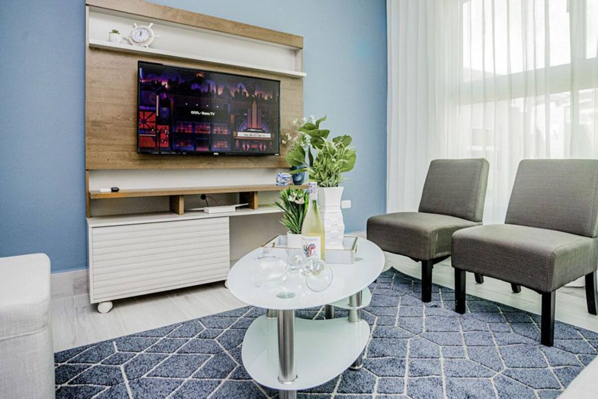 A modern living room with a flat-screen TV mounted on the wall, two grey armchairs, a white coffee table with a vase and glasses, and a patterned rug.