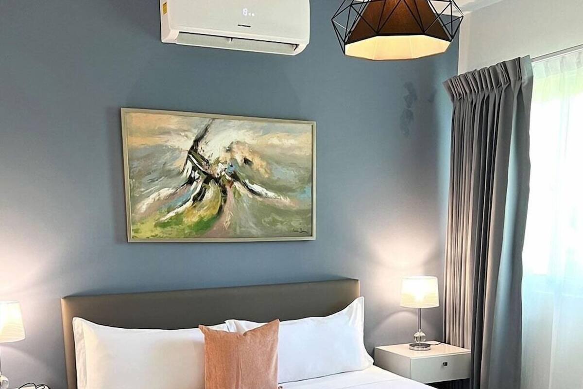 A hotel room with a bed, a painting, an air conditioner, a lamp, and a window with curtains.