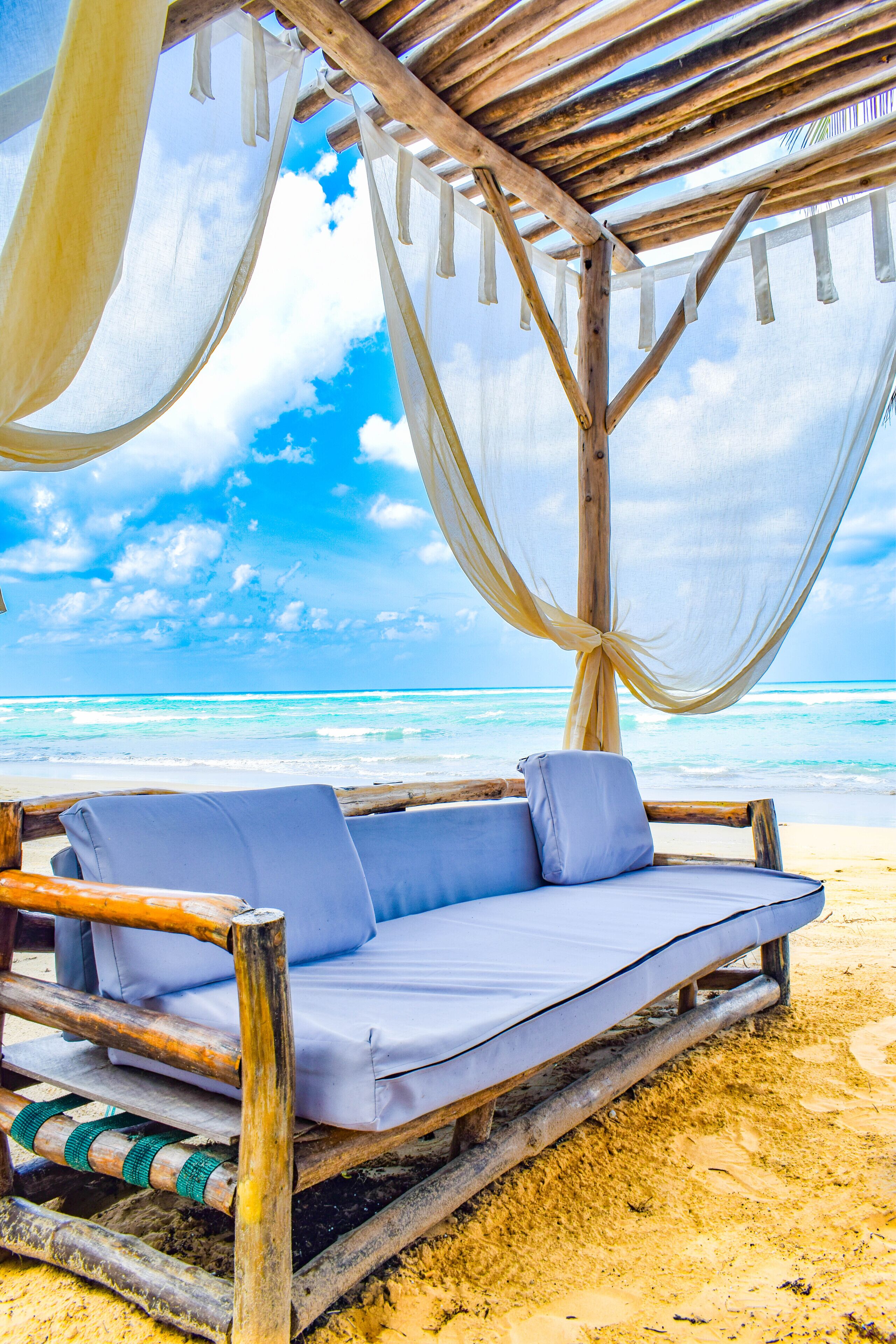 Une chaise de plage en bois, dotée d’un coussin, est placée sur une plage de sable, offrant une vue dégagée sur l’océan, et une structure en bois la surplombe.