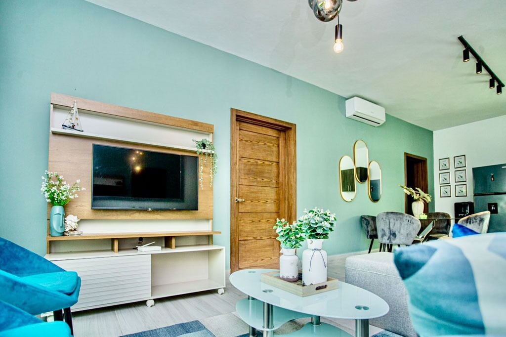 A modern living room with a large flat-screen TV mounted on the wall, a wooden door, and a round glass coffee table with decorative items.