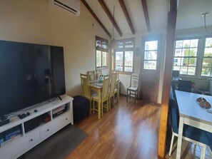 Ein Zimmer mit Fernseher, Esstisch aus Holz, Stühlen und einem Fenster mit Blick auf Gebäude.