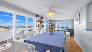 Un espace repas comprenant une table, des chaises, un ventilateur de plafond et de grandes fenêtres donnant sur la mer.