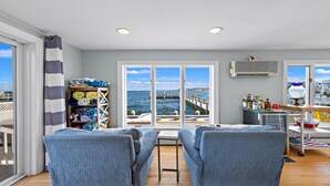 Une pièce avec vue sur la mer, une étagère en bois où sont pliés des vêtements, une petite table avec des bouteilles et deux chaises bleues.