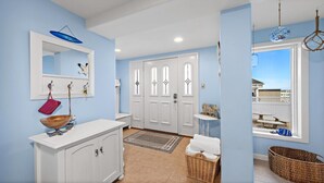Une salle de bain avec un meuble-lavabo blanc, un miroir, un bol en bois, un panier en osier et une vue sur la plage par une fenêtre.