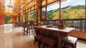 A spacious room with a large wooden table and benches, large windows with a view of rolling hills, and a wooden ceiling with exposed beams. - Longji Rice Terraces Heshe Homestay (Longsheng)