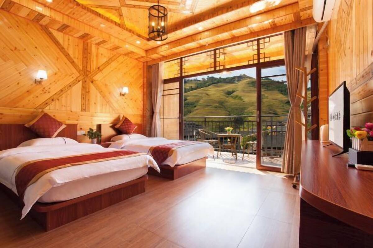 A wooden interior with two beds, a desk, and a balcony with a view.