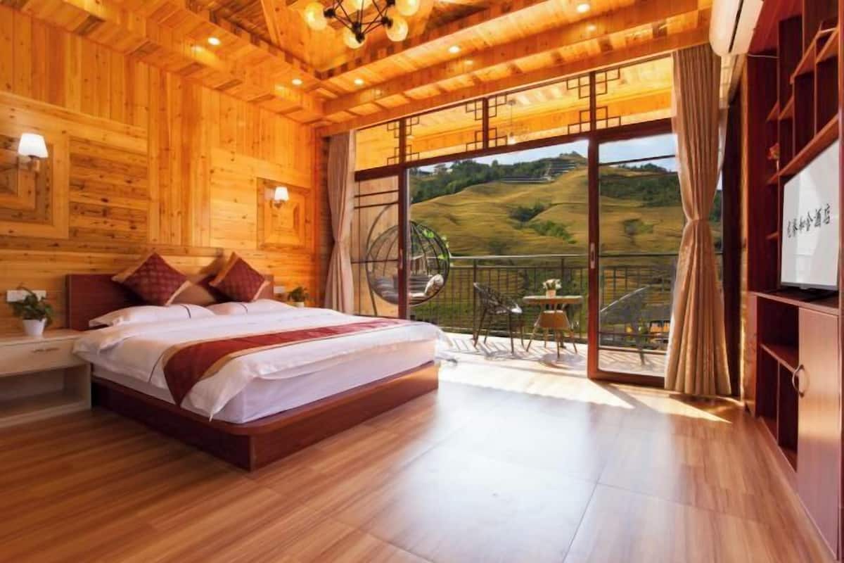A wooden interior bedroom with a bed, a small table, and a view of a green landscape through a glass door.