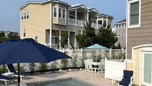 Una piscina con sillones de descanso y sombrillas, rodeada por una valla y un edificio con varios balcones.