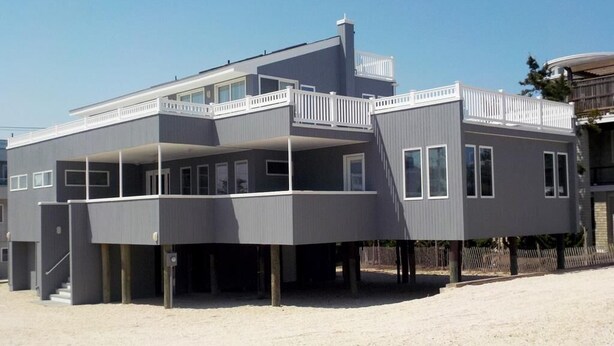 A modern two-story house with a balcony, a covered entryway, and a sandy ground. - OCEAN VIEWS! BEAUTIFUL HOME SLEEPS 14! (North Beach)