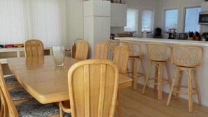 A dining area with a wooden table and wicker chairs, a kitchen with white cabinets, and a window with blinds. - OCEAN VIEWS! BEAUTIFUL HOME SLEEPS 14! (North Beach)