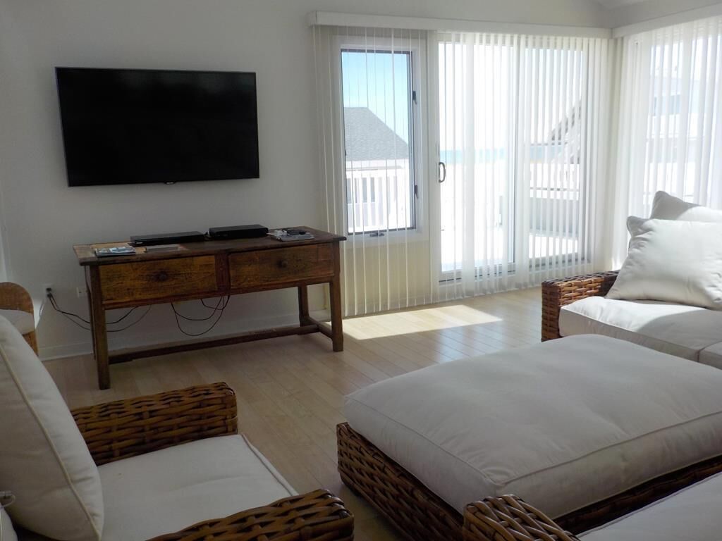 A hotel room with a flat-screen TV mounted on the wall, a wooden console table with electronic devices, a large bed with white bedding, wicker furniture, and a sliding glass door leading to a balcony.