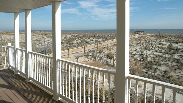 Une véranda couverte, dotée de balustrades blanches, offre une vue sur une plage bordée de dunes et un ciel dégagé.