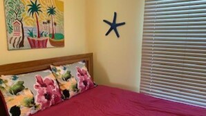 Une chambre avec une couvre-lit rouge, une tête de lit en bois et un mur orné d’un tableau représentant un paysage tropical et d’une décoration en forme d’étoile.