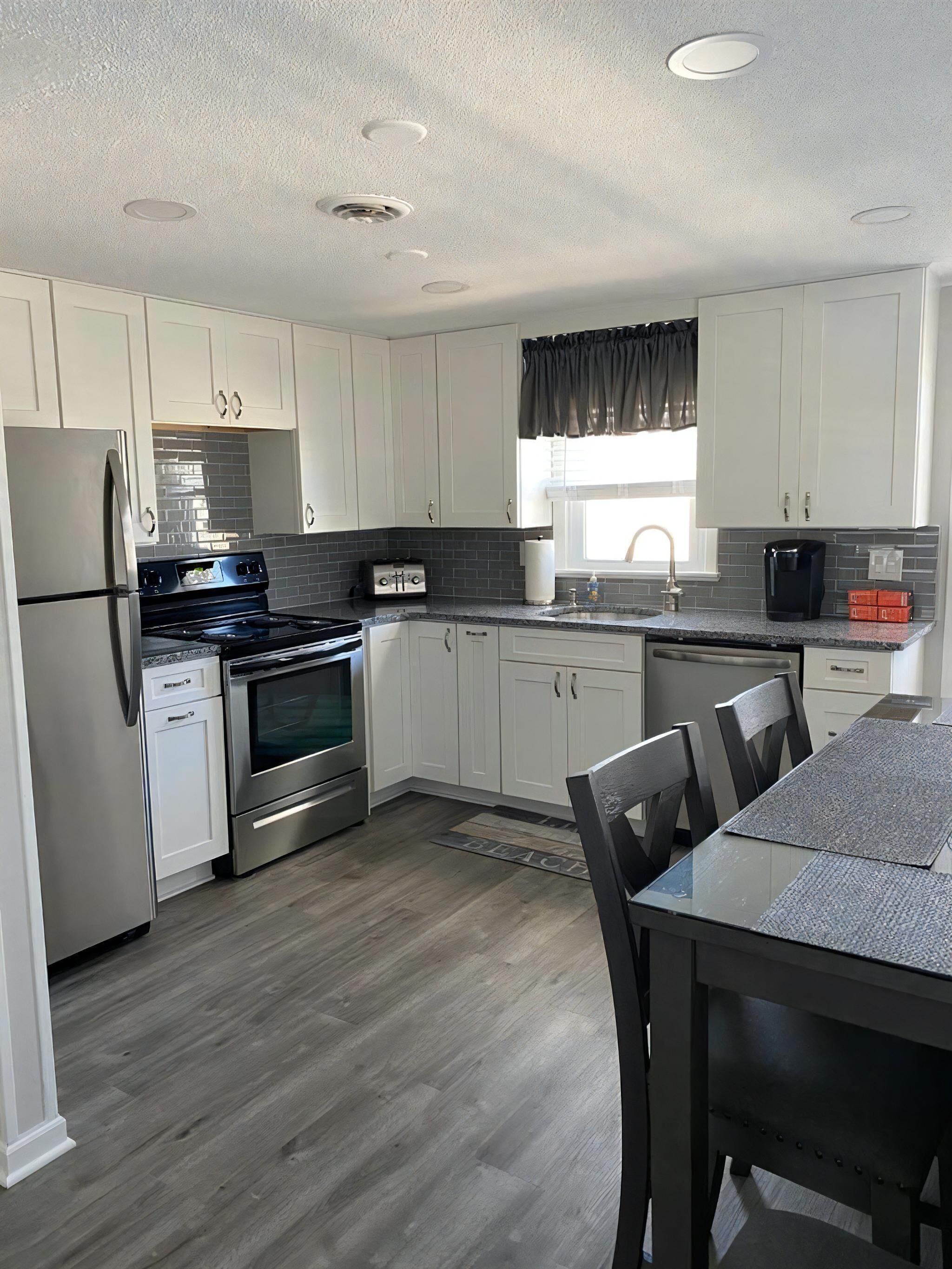 A modern kitchen with white cabinets, stainless steel appliances, a dining area with a glass table, and a window with a view.