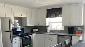 A modern kitchen with white cabinets, stainless steel appliances, a dining area with a glass table, and a window with a view. - Newly renovated duplex located on the ocean block in the heart of Surf City. (Surf City)