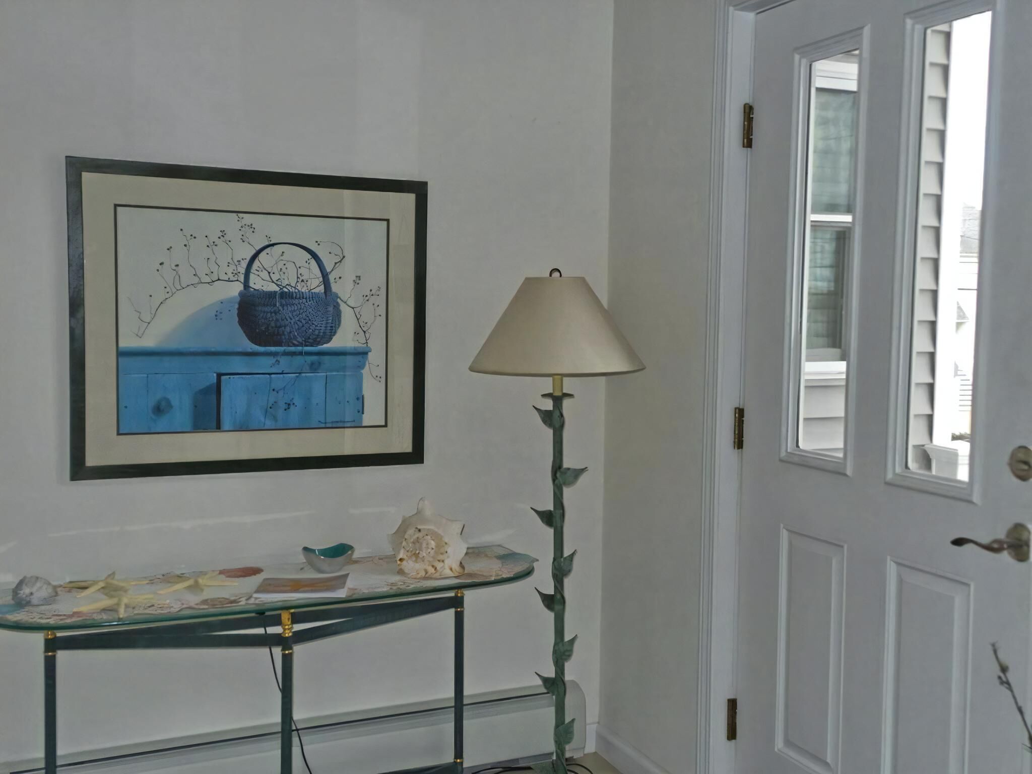 Une pièce avec une œuvre d’art encadrée au mur, une table ornée d’objets décoratifs, une lampe et une porte avec une fenêtre.