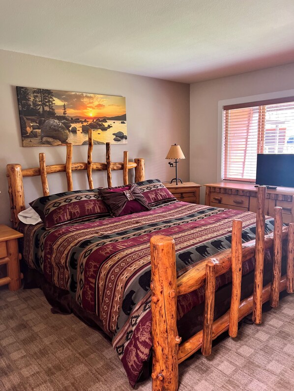 A bedroom with a wooden bed frame, a patterned bedspread, a painting of a sunset, a lamp, and a television on a wooden stand.