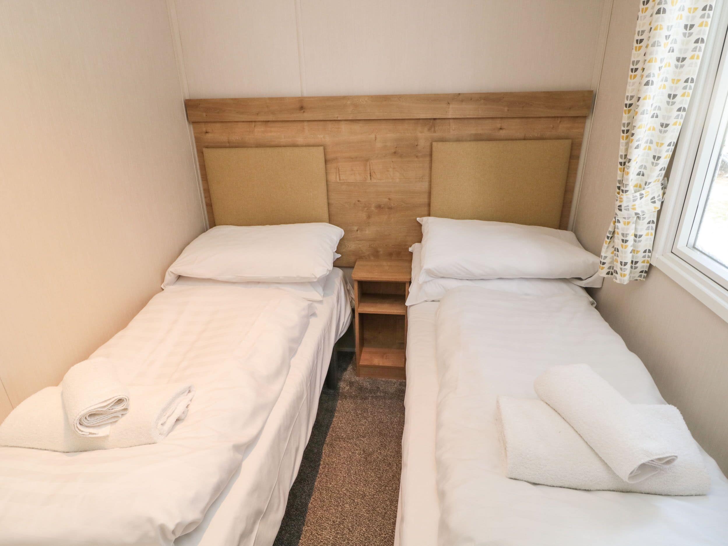 Quarto duplo com duas camas de solteiro, cabeceira de madeira, roupa de cama branca e janela com cortinas estampadas.