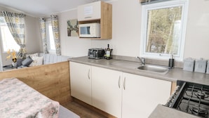 A compact kitchen with a microwave, toaster, and sink. There is a window with a view of trees and a sofa with a floral cover in the adjacent living area. - Daisy's Retreat (Morpeth)