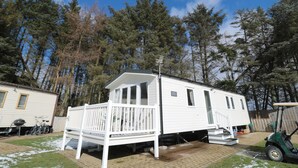 A white mobile home with a porch and a small deck, surrounded by trees and a golf cart. - Daisy's Retreat (Morpeth)