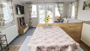 A cozy room with a wooden bench, a table with a floral tablecloth, a TV, and a window with curtains. - Daisy's Retreat (Morpeth)