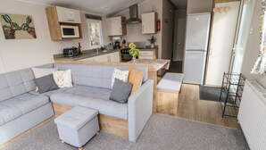 A modern living room with a grey sectional sofa, a small ottoman, and a kitchen area with a microwave, stove, and refrigerator. - Daisy's Retreat (Morpeth)