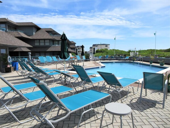A poolside area with blue lounge chairs and umbrellas, a clear sky, and buildings in the background.