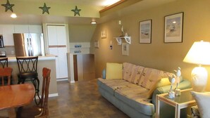 A living room with a sofa, a dining table with chairs, a lamp, and a kitchen area in the background.
