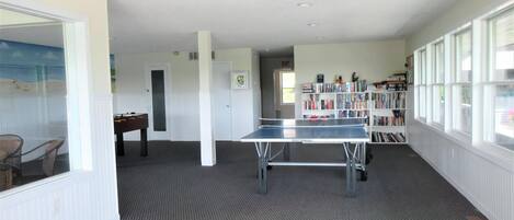 A room with a ping pong table, a bookshelf, and a view through a window.