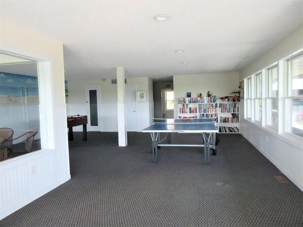 A room with a ping pong table, a bookshelf, and a view through a window.