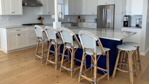 A modern kitchen with white cabinetry, a central island with bar stools, a stainless steel refrigerator, and a hanging woven light fixture. - Surf City Oceanfront With Panoramic Views (Surf City)