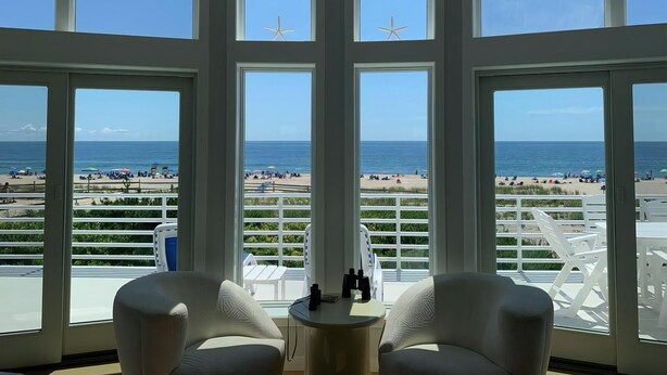 A room with large windows, a white chair, a small table, and a view of the beach. - Surf City Oceanfront With Panoramic Views (Surf City)