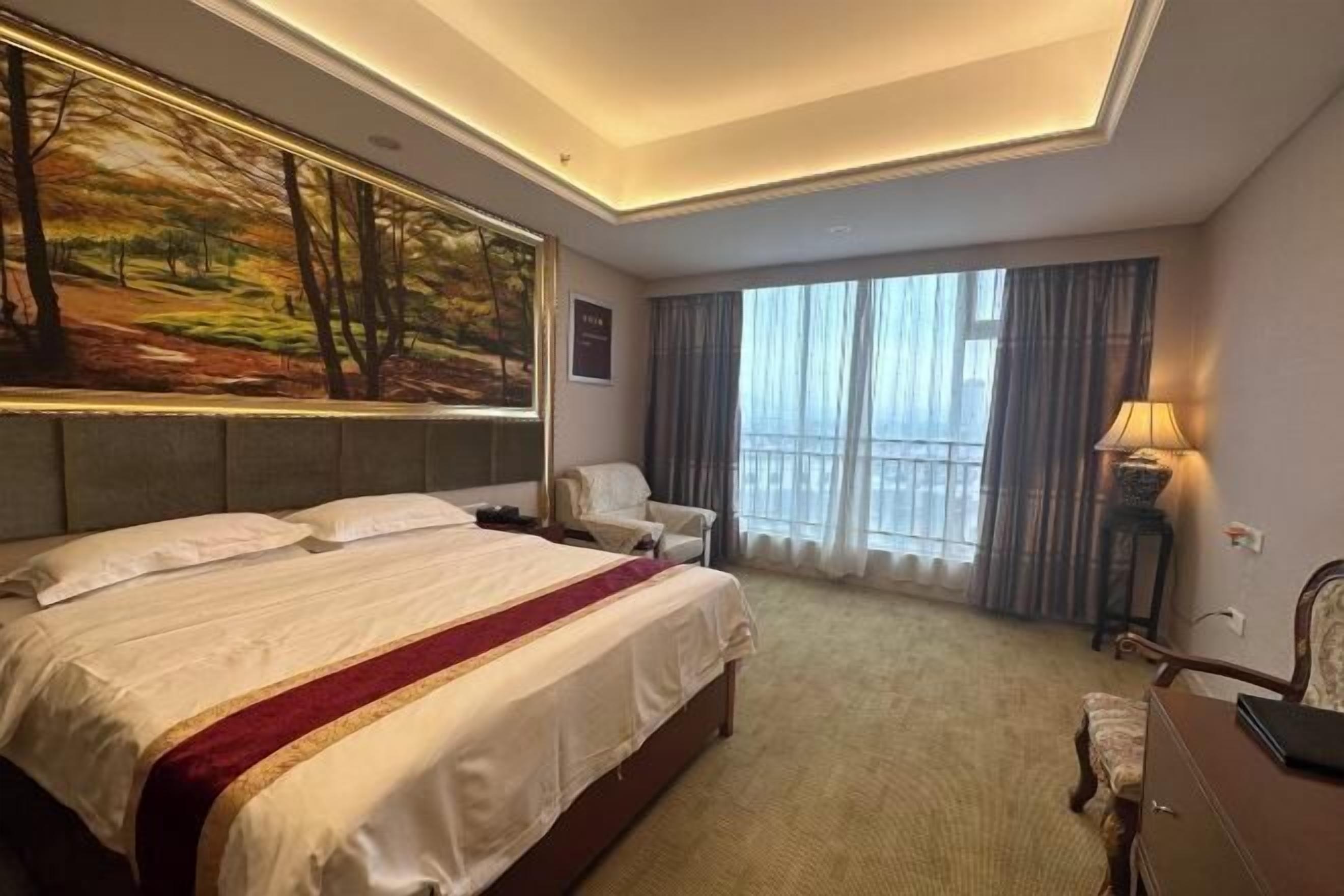 Een hotelkamer met een groot bed, een bureau, een stoel, een schilderij aan de muur en een raam met gordijnen.