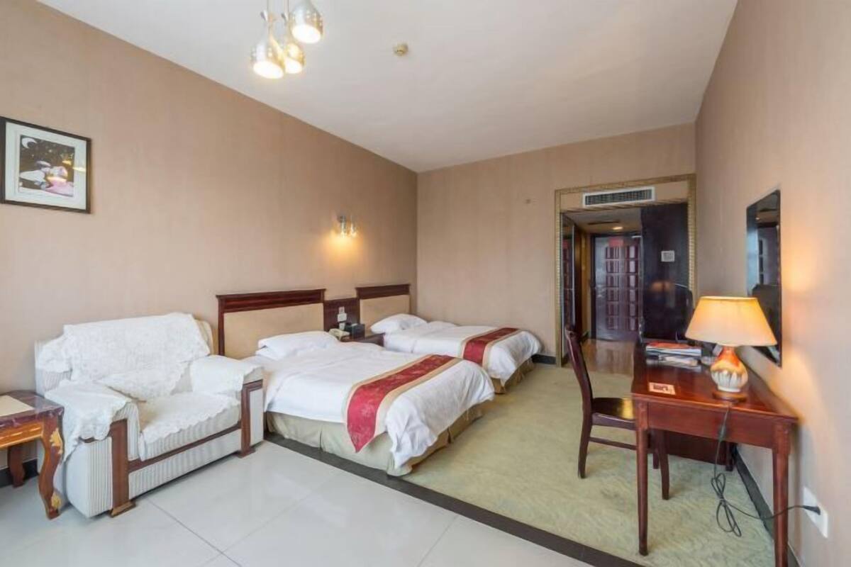 A hotel room with two beds, a sofa, a desk, and a chair. There is a door leading to another room and a wall-mounted TV.