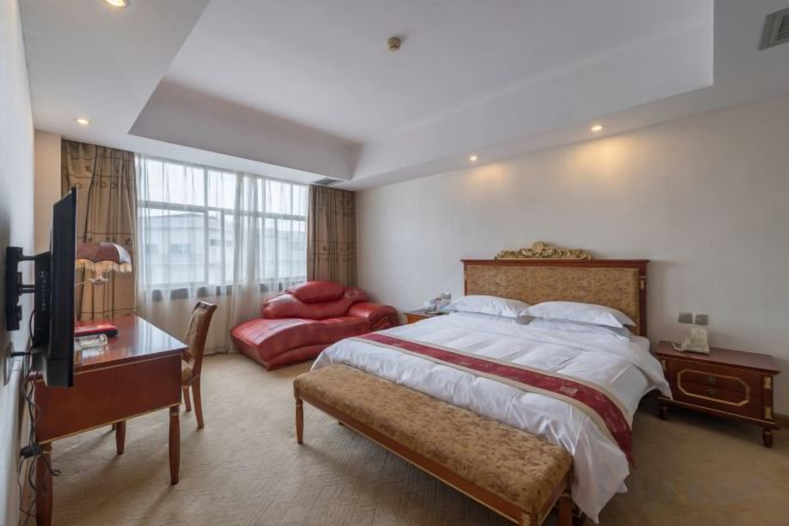 A hotel room with a bed, a desk with a chair, a red sofa, and a television.